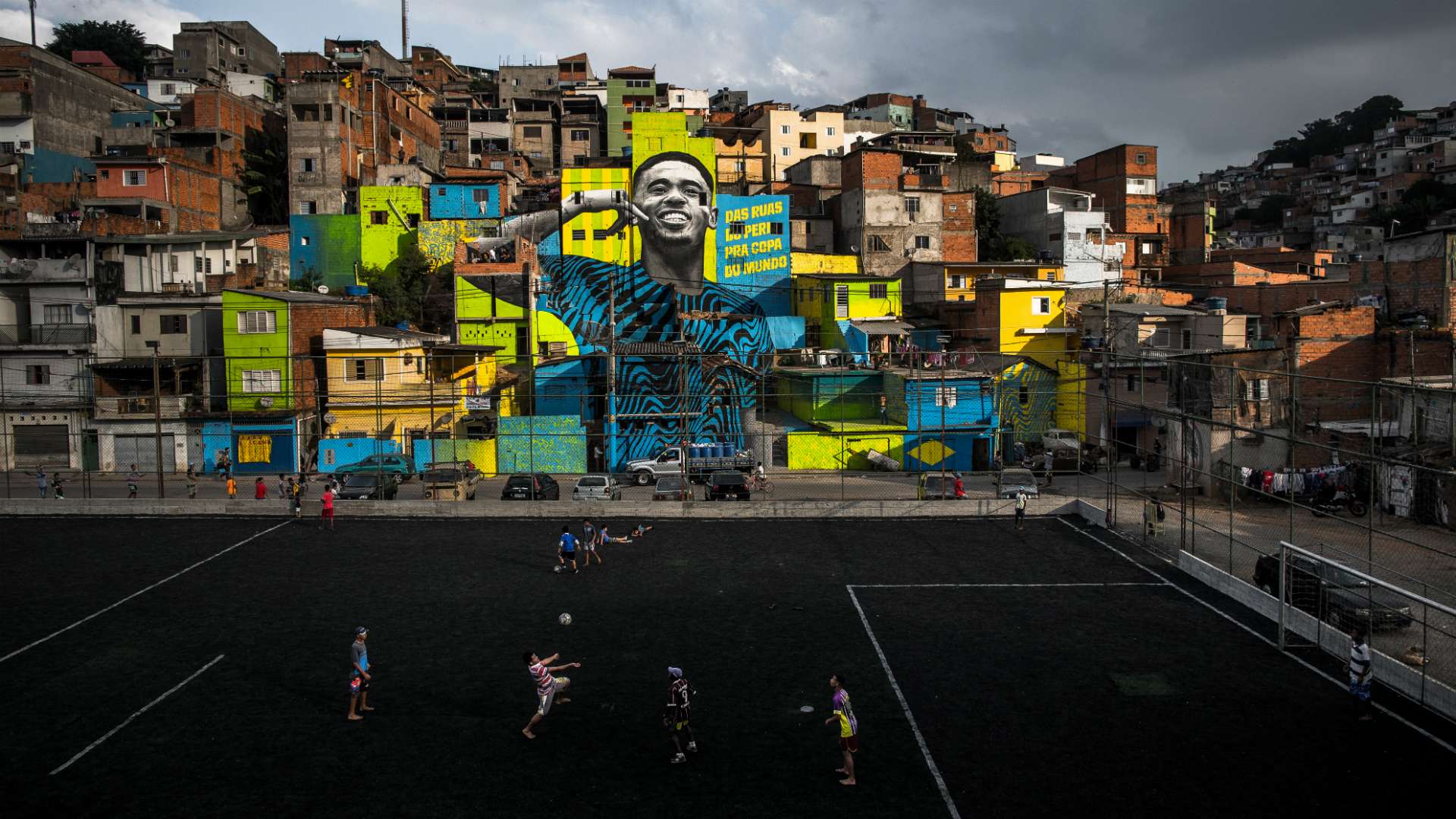 Il murale di un calciatore su un quartiere colorato in collina, con dei ragazzi che giocano a pallone su un campo in primo piano